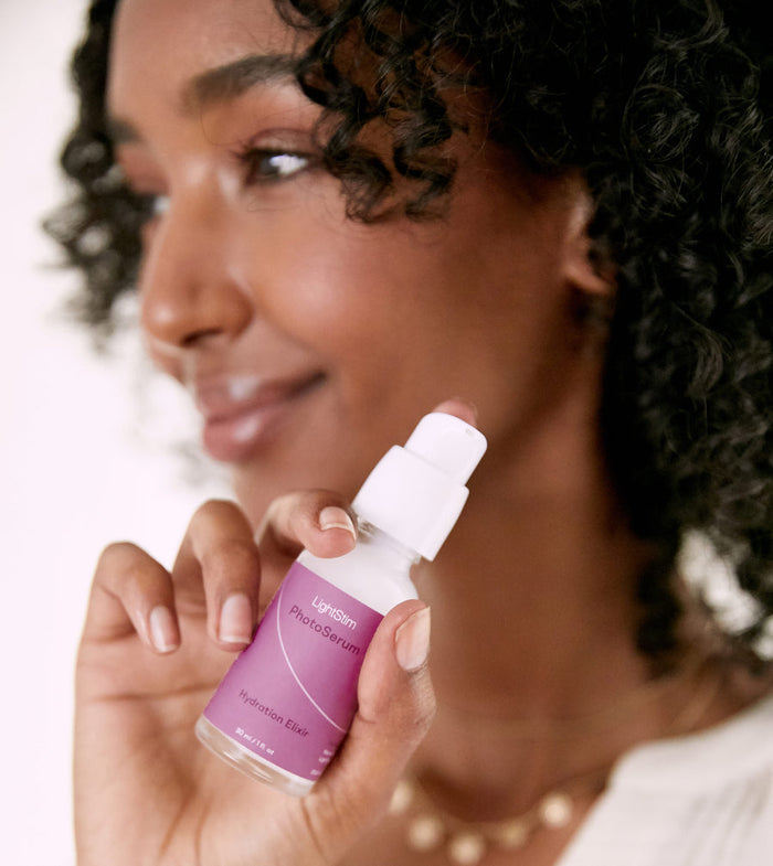 Woman with curly hair holding a bottle of LightStim PhotoSerum | LightStim, a skincare product in a purple-labeled bottle, close to her face with a soft smile.