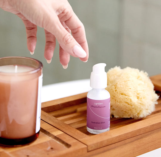 Hand reaching for a bottle of LightStim PhotoSerum | LightStim on a wooden tray next to a candle and natural bath sponge in a clean, spa-like setting.